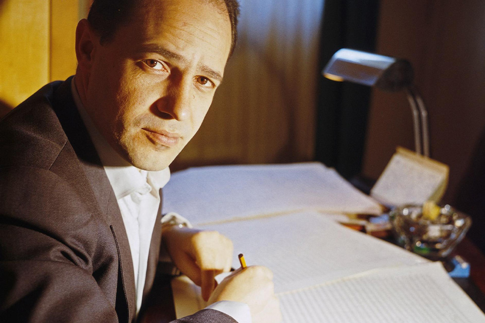 Pierre Boulez wears a suit and holds a pencil to may layers of manuscript paper
