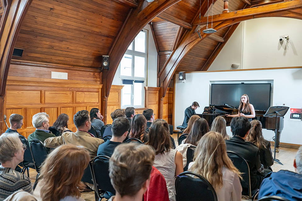 A singer and pianist perform to an audience in a brightly lit room with wooden walls